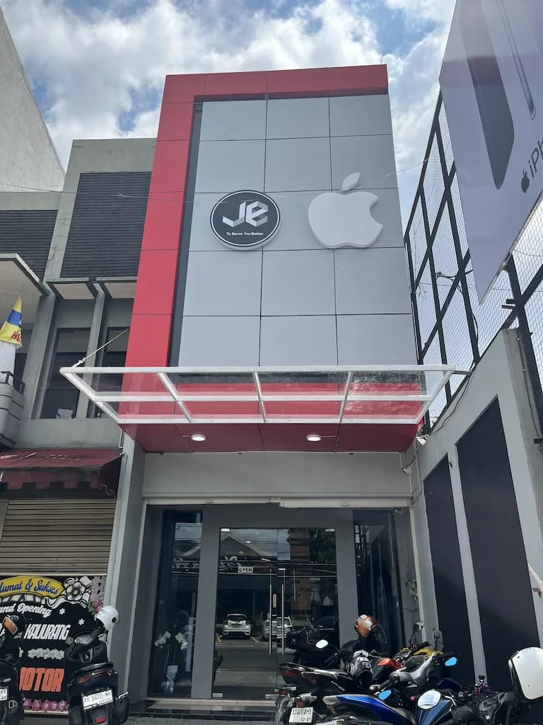 Fasad toko JE merah-abu dengan logo Apple menyala dan kanopi kaca